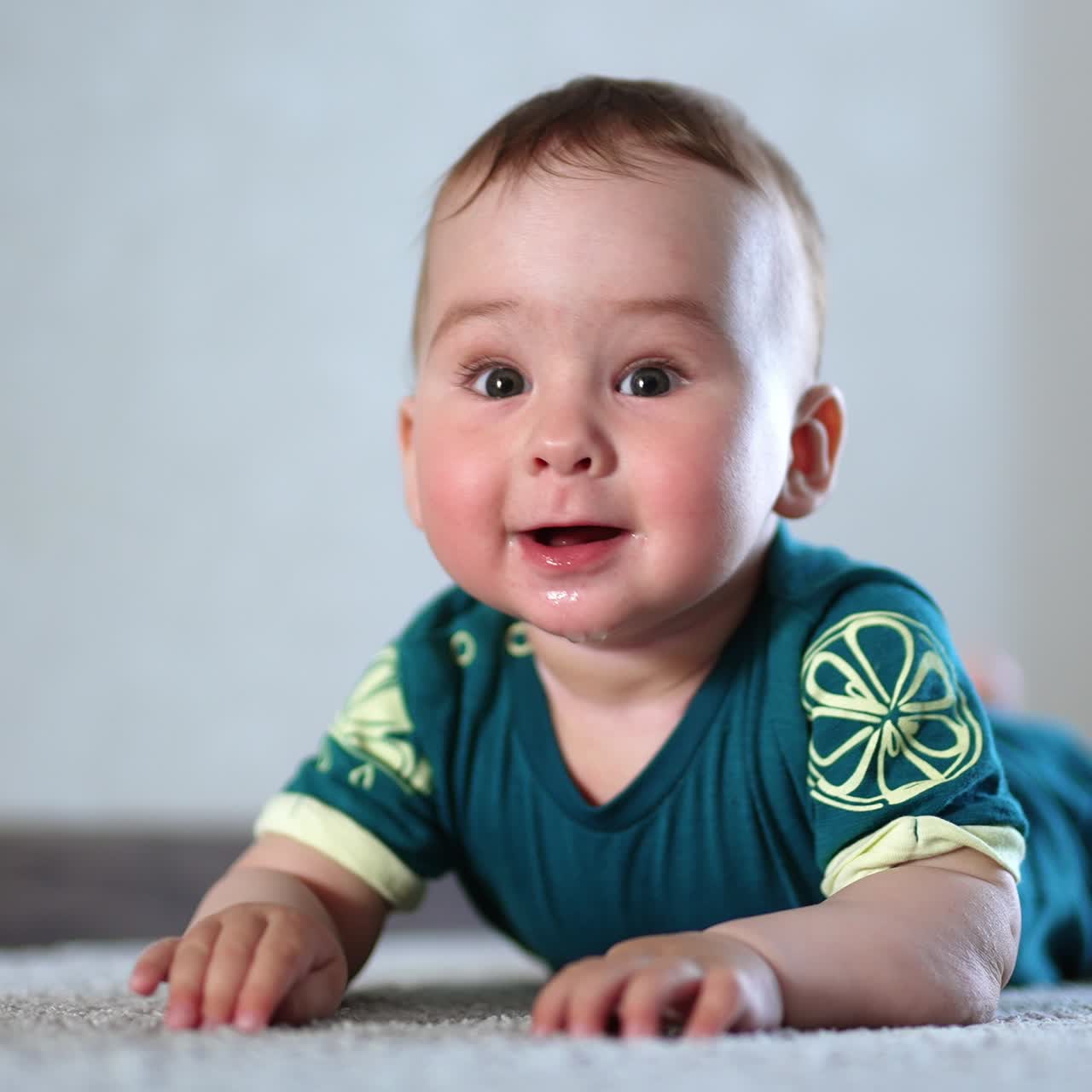 Happy smiling infant boy lies on belly on the floor. Active baby claps his hands and feet over the carpet. Light backdrop