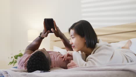 Happy-diverse-couple-using-smartphone-and-lying-in-bedroom