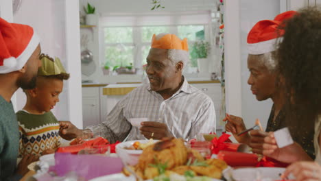 Familia-Multigeneracional-Con-Sombreros-De-Papel-Leyendo-Chistes-De-Galletas-Navideñas-Mientras-Comen-Juntos