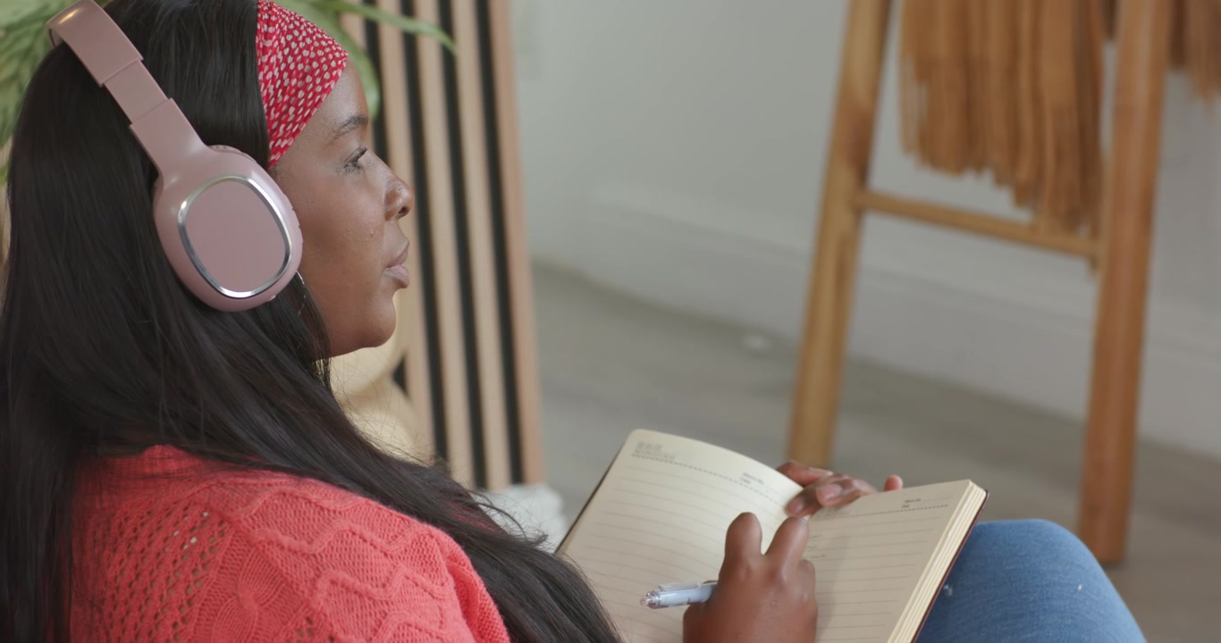 African American woman listening on headphones, writing, tapping notes, glancing up