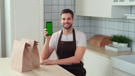 un hombre atractivo sostiene un teléfono inteligente con una pantalla verde en la mano y sonríe mientras está sentado en la cocina.