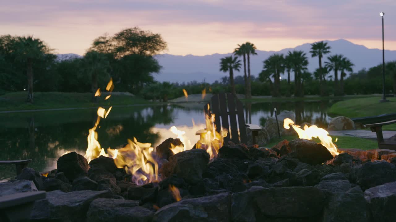 Tall flames provide plenty of warmth from a gas-burning campfire on a tranquil lake, while the background sky is beautifully colored by the setting sun. Slowly tilt slow motion shot