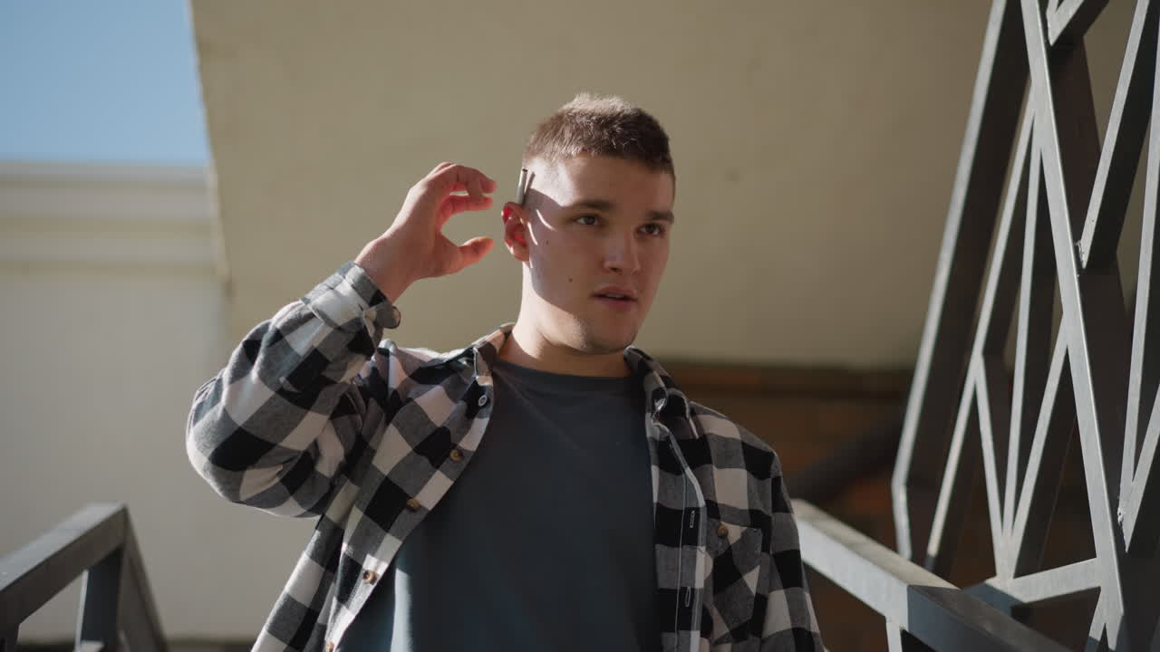 man in black white check flannel shirt walking down outdoor metal stairs against beige wall removes cigarette from ear flicks lighter holds flame near cigarette tip prepares inhale
