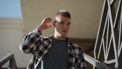 man in black white check flannel shirt walking down outdoor metal stairs against beige wall removes cigarette from ear flicks lighter holds flame near cigarette tip prepares inhale