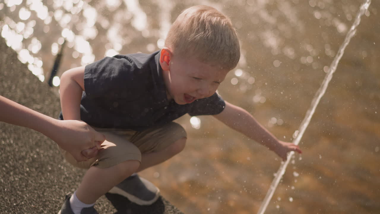 Niño alegre toca un chorro de agua de la mano de su madre en una plaza.