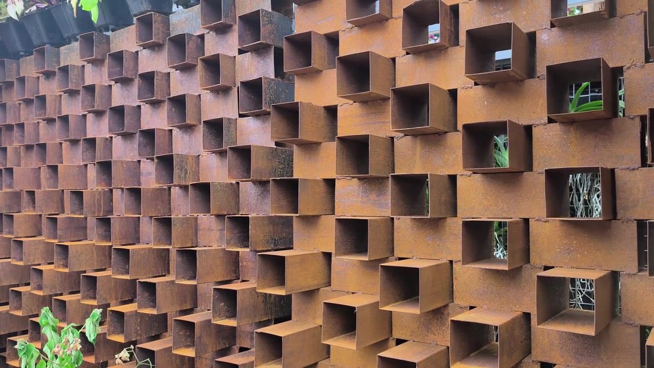 Una grande parete di ventilazione è realizzata con sezioni cave rettangolari corte in acciaio saldate insieme. La finitura in metallo arrugginito aggiunge un'estetica rustica distintiva alla struttura