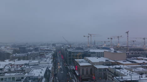 Vista-Aérea-De-Edificios-Nevados-En-Una-Gran-Ciudad.-Grupo-De-Grúas-Torre-En-El-Sitio-De-Construcción.-Coches-Circulando-Por-La-Calle-Al-Atardecer.-Berlín,-Alemania