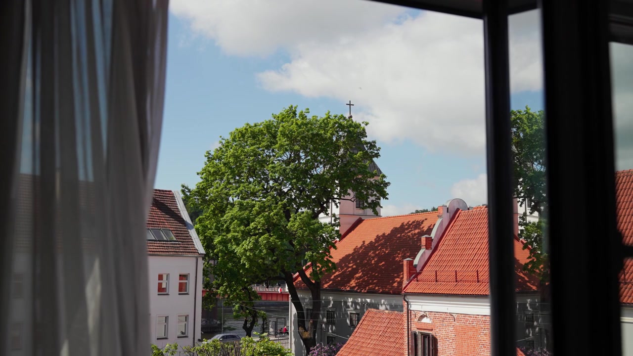 Ruhige Aufnahme von innen, Blick aus einem offenen Fenster auf rot gedeckte Dächer, belaubte Bäume und eine Kirche mit Kreuz bei hellem Tageslicht, die eine ruhige, luftige Stimmung erzeugt