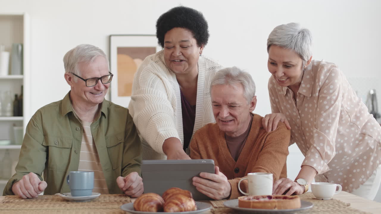 Medium shot of cheerful diverse multiethnic seniors smiling, having fun conversation, looking at tablet computer at table
