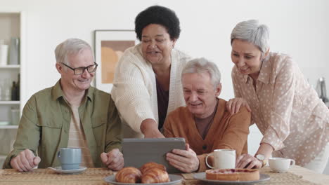 medium shot of cheerful diverse multiethnic seniors smiling, having fun conversation, looking at tablet computer at table
