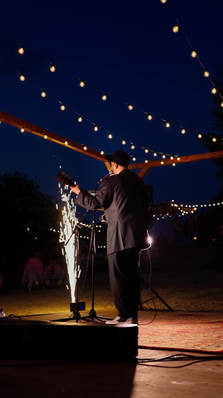 Musician Performing Live on Outdoor Stage with Sparkler and String Lights at Night