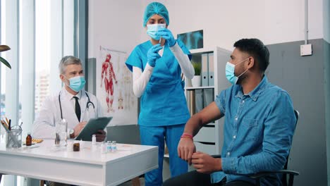 Joven-Hindú-Enfermera-Especialista-En-Atención-Médica-Que-Inyecta-La-Vacuna-Covid-19-A-Un-Apuesto-Paciente-Masculino-En-El-Gabinete-De-La-Clínica-Con-Máscara-Mientras-El-Médico-Principal-Lee-El-Análisis,-La-Vacunación-Contra-El-Coronavirus