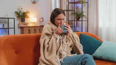 young unhealthy sick ill woman covered by warm blanket drinks coffee freezes from cold flu at home