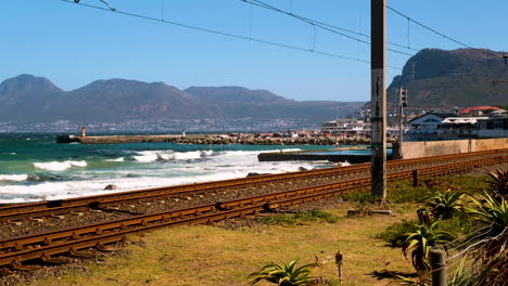 View-of-Kalk-Bay-harbour-from-over-old-railway-tracks,-waves-breaking-on-shore