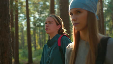 friends walk through a lush green forest, both wearing relaxed expressions, one wears a blue hair tie and carries a backpack, while the other drapes a cloth over her bag with her hair tied back