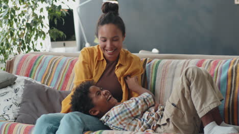 Joyous-Mother-Tickling-Excited-Kid-on-Sofa-at-Home