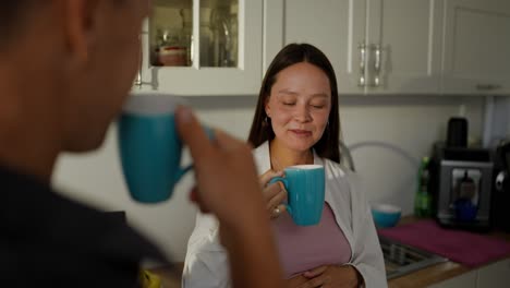 Over-the-shoulder-a-happy-pregnant-brunette-woman-drinking-tea-from-a-blue-cup-and-chatting-with-her-husband-at-home-in-the-morning