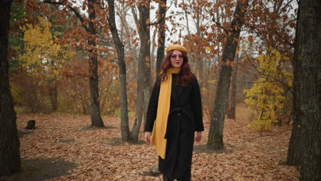 young tourist in black coat, yellow beret, and muffler touches a dry branch and looks around with excitement in an autumn-covered forest