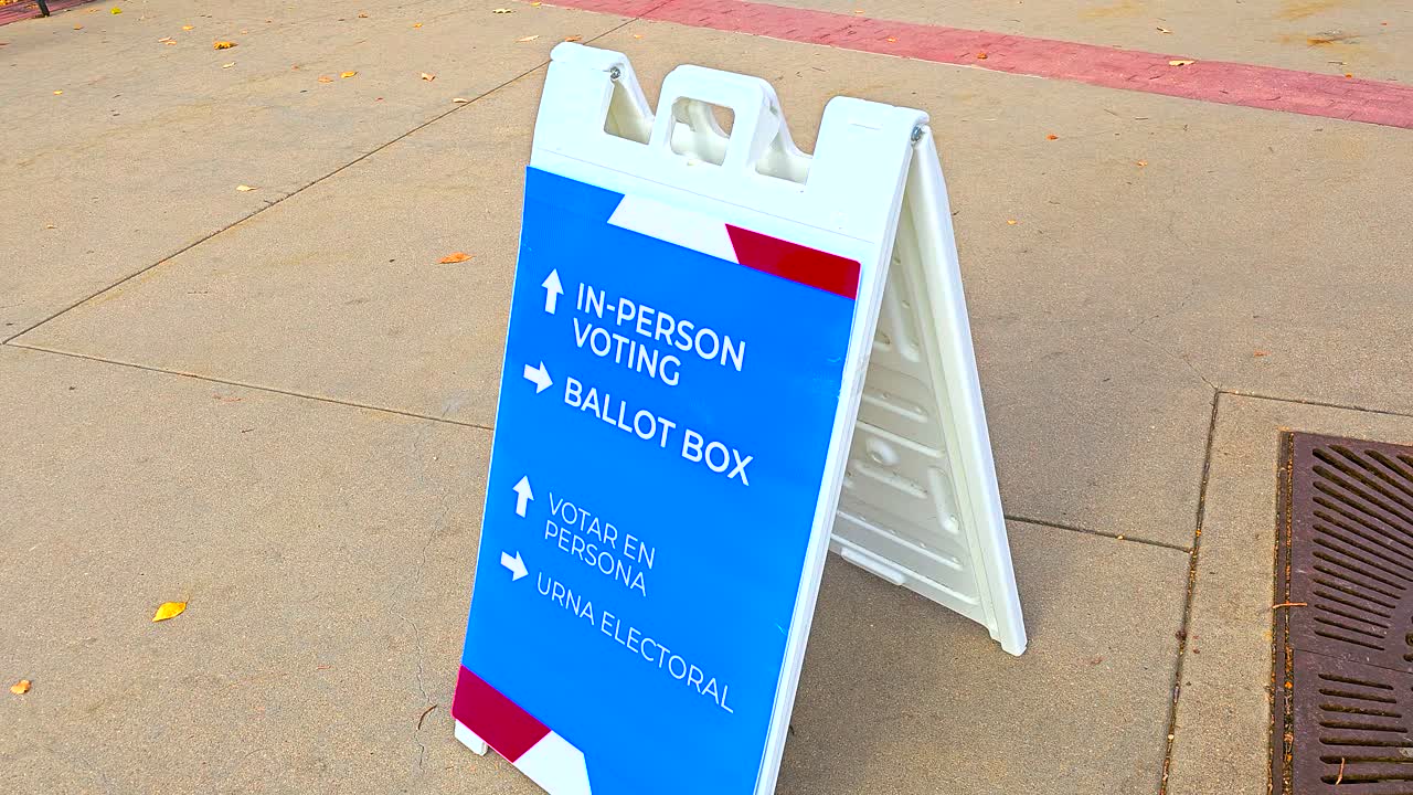 A ballot box sign with large arrows directs citizens to the polling place, with Spanish text Votar en Persona and Urna Electoral emphasizing civic duty, democracy, accessibility, equal participation