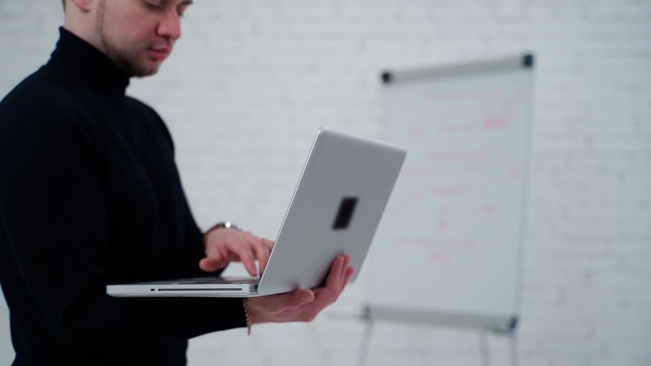 Handsome young man working on a laptop. Man in black sweater standing in the light studio and talking to someone through the internet on a computer.