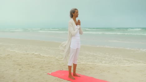 Side-view-of-active-senior-African-American-woman-doing-yoga-on-exercise-mat-at-the-beach-4k