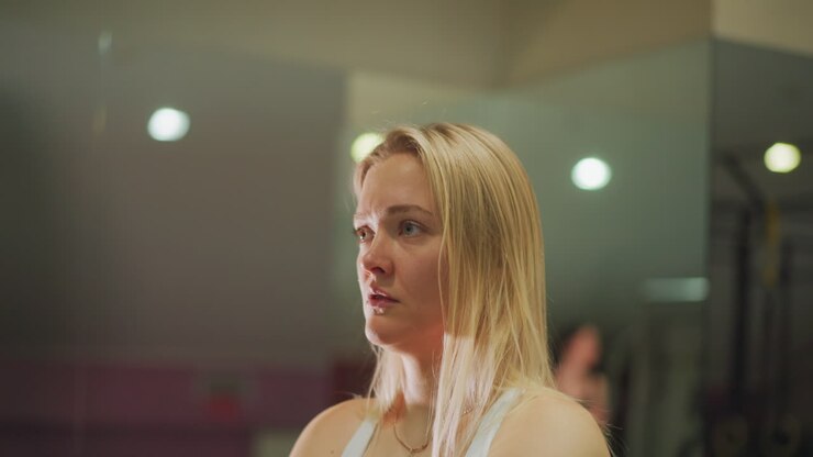 Donna bionda in palestra che osserva la lezione, luci soffuse da studio e sagome sfocate del gruppo, espressione concentrata e tensione silenziosa, atleta e allieva che si preparano mentalmente, un sottile ritratto di determinazione.