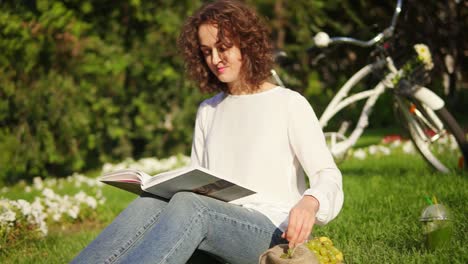 Young-woman-in-white-shirt-and-blue-jeans-is-reading-a-book-sitting-on-the-grass-in-park-and-taking-grapes.-Her-city-bicycle