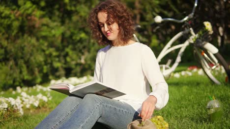 una mujer joven con camisa blanca y vaqueros azules está leyendo un libro sentado en la hierba en el parque y tomando uvas. su bicicleta de la ciudad