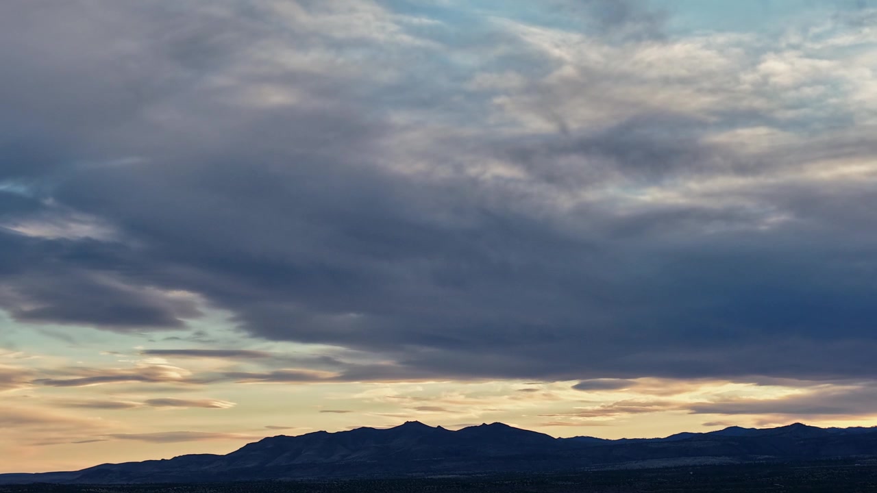 A cinematic 4k drone timelapse captures moody sunset clouds and shifting light over a silhouetted mountain ridge. This dramatic atmospheric transition provides an epic and scenic nature background.