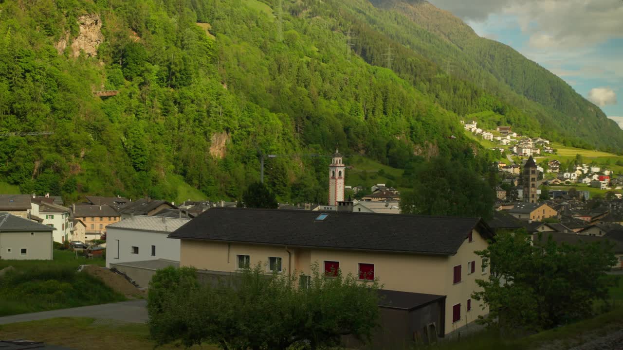 Pohled z výšky na domy a kostelní věž v Poschiavu s bujně zeleným úbočím hory v pozadí. Krásná architektura švýcarské vesnice a tradiční alpská osada v regionu Graubünden.