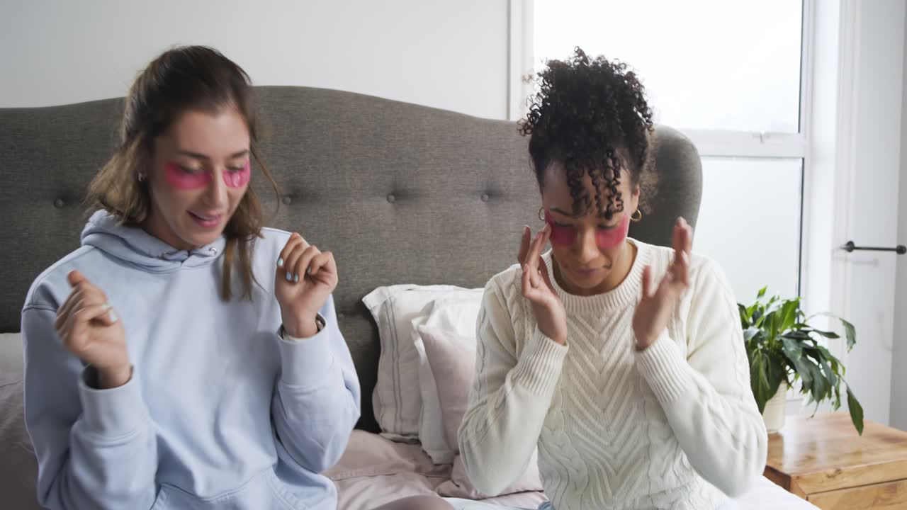 Diverse female friends tapping phone on bed and applying pink eye patches for skincare selfies