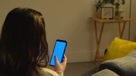 rear view of young woman spending evening at home sitting on sofa looking at blue screen mobile phone 2