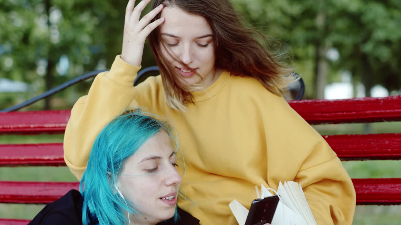 공원의 벤치에 휴식을 취하는 레즈비언 커플, 책에 손을  젊은 여성, 여자친구의 염색 된 머리카락을 만지고, 공원의 치에서 휴식을 취하면서 음악을 듣고 소셜 미디어를 탐색합니다.