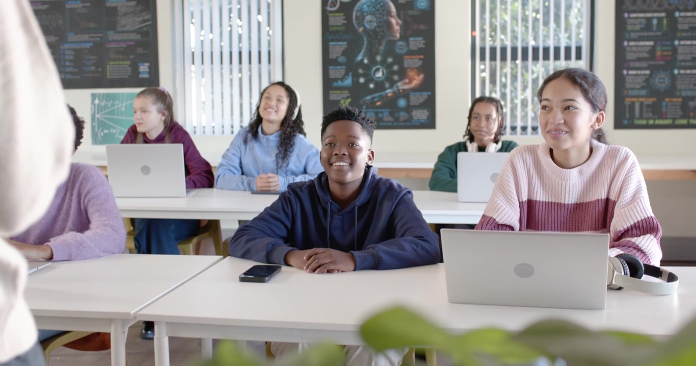 Colegas de classe adolescentes na sala de aula virando-se para o professor entrando pela esquerda, sorrindo, verificando laptops