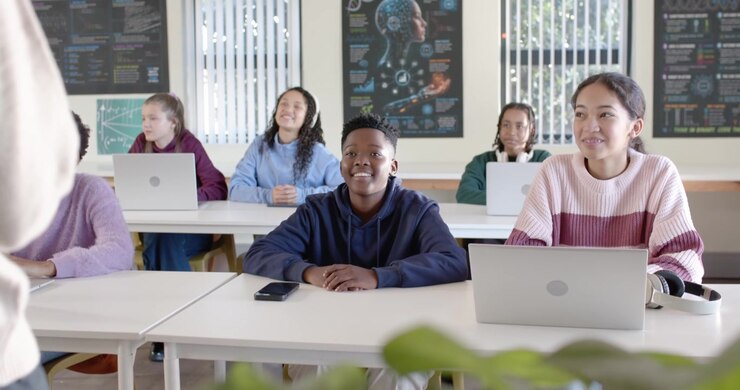Teen classmates in classroom turning toward teacher entering from left, smiling, checking laptops