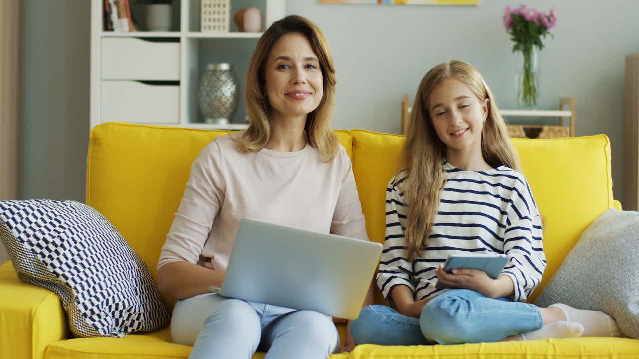 Blonde Mother And Daughter Sitting On Yellow Couch While They Using Laptop Computer And Tablet And Looking At Camera In The Living Room