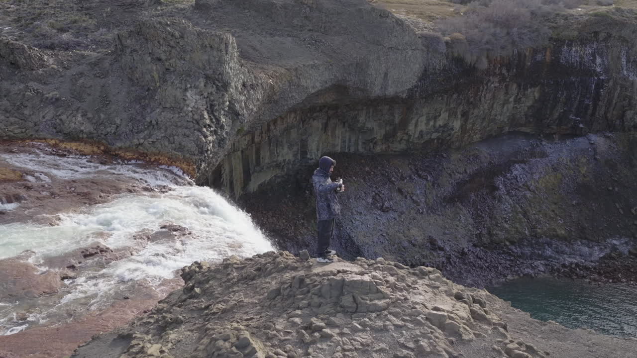 Slow drone orbit of a traveler standing on the brink of Agrio Waterfall near Caviahue, Neuquén, Argentina, with foaming river, columnar basalt, and teal plunge pool set in a windswept volcanic steppe