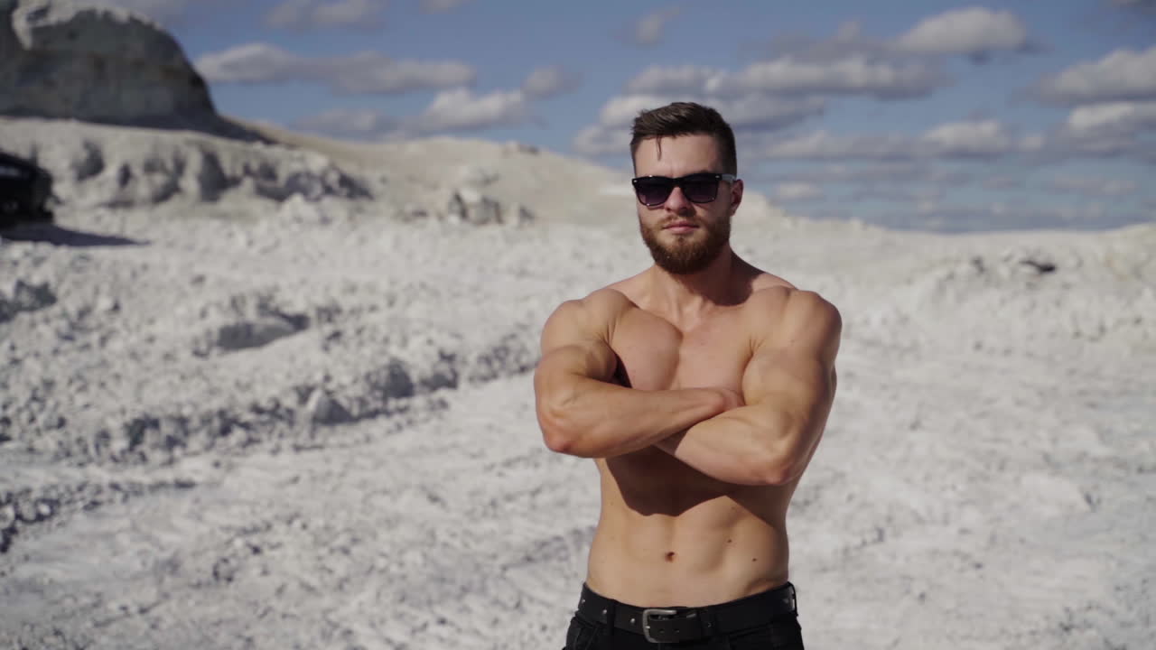 Portrait of shirtless athlete. Handsome guy with beard in sunglasses with muscular body posing on white canyon in summer. Camera motion around.