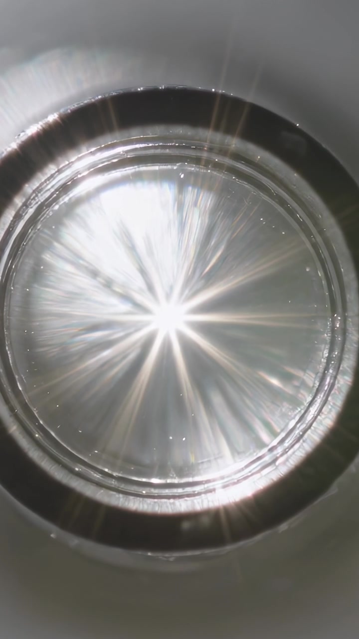 Video vertical: Luz cambiante incidiendo en una lente con montura metálica sobre un banco, reflejando rayos de luz en forma de estrella y motas.