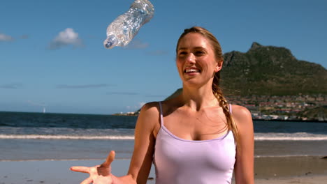 rubia en forma sonriendo y arrojando su botella en la playa