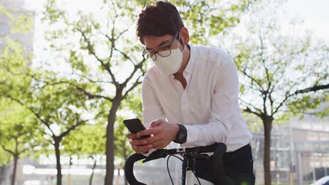 Hombre-Asiático-Con-Mascarilla-Usando-Un-Teléfono-Inteligente-Mientras-Se-Apoya-En-Su-Bicicleta-En-La-Calle