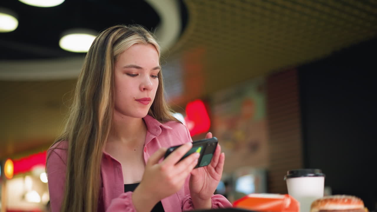 mujer de vestido rosa se centra en su teléfono, posiblemente tomando una foto de algo delante de ella, taza de café y hamburguesa en la mesa en primer plano, mientras que el fondo está suavemente borroso