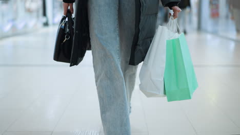 lady in jeans trousers and black jacket walking in well-lit retail space carrying black handbag and two shopping bags, one white and one mint green, blurred background with other shoppers