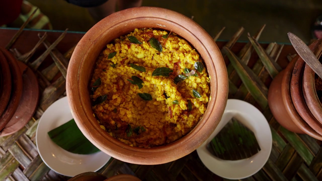 Vista dall'alto di una mano che serve curry di lenticchie giallo curcuma da una pentola di terracotta con un cucchiaio di legno in un ambiente tradizionale di pasto in un villaggio dello Sri Lanka