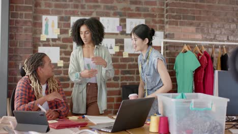 diseñadores diversos felices usando portátiles y discutiendo el trabajo en el estudio de diseño de moda en cámara lenta