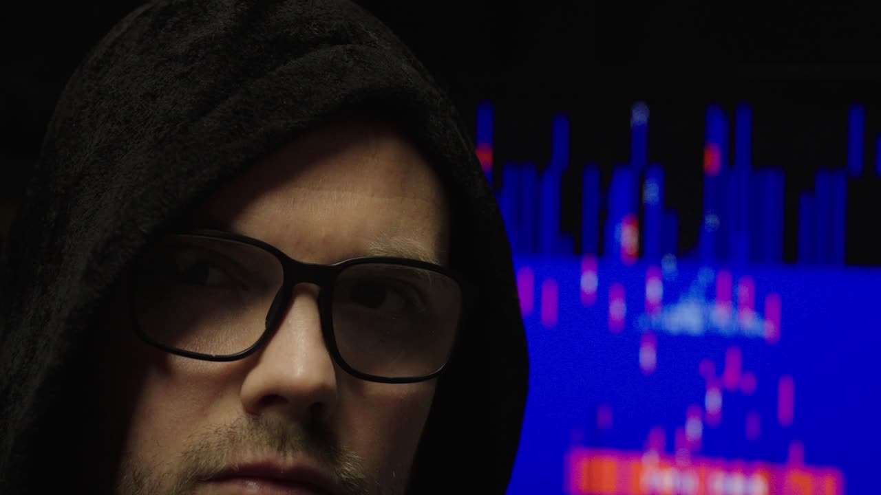 Primo piano suggestivo di un uomo incappucciato con gli occhiali davanti a uno schermo di computer sfocato con codice e messaggio di avviso, a rappresentare minacce alla sicurezza informatica e hacking anonimo