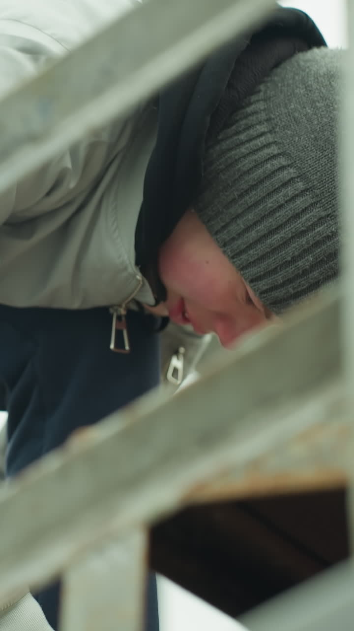 Close-up of a young boy wearing a gray jacket, blue joggers, and a green beanie, focused on what he is doing near an iron railing