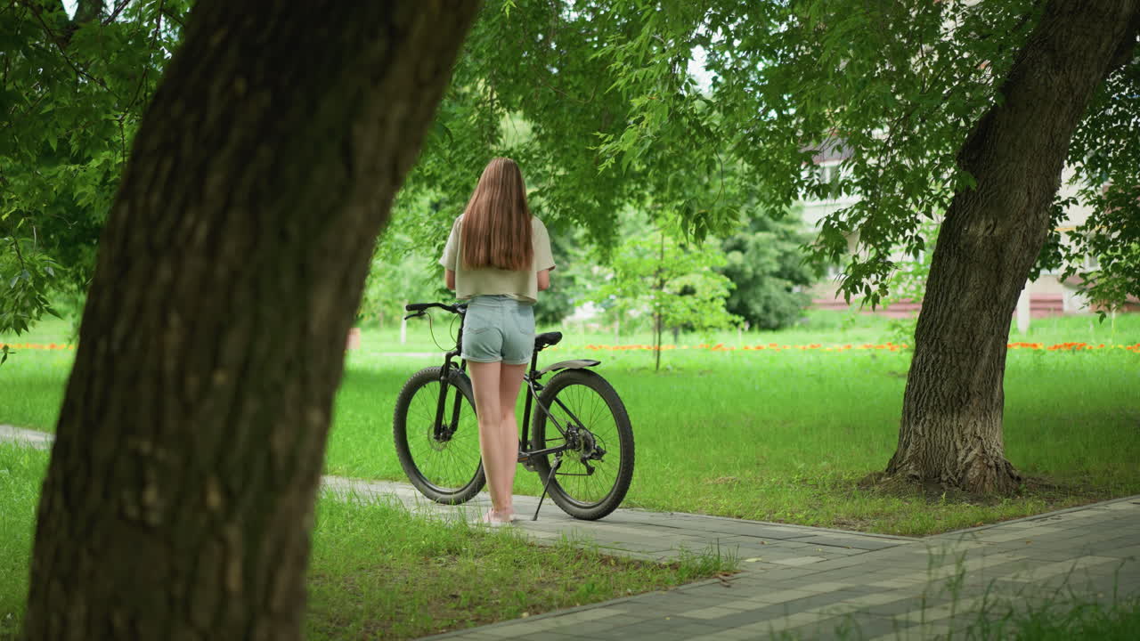 분홍색 스니커와 청바지 쇼트를 입은 젊은 여성이 자전거 좌석에 물건을 놓고 자전거 길에서 자전거로 걸어가고, 배경에는 울창한 나무, 초록색, 건물이 있습니다.