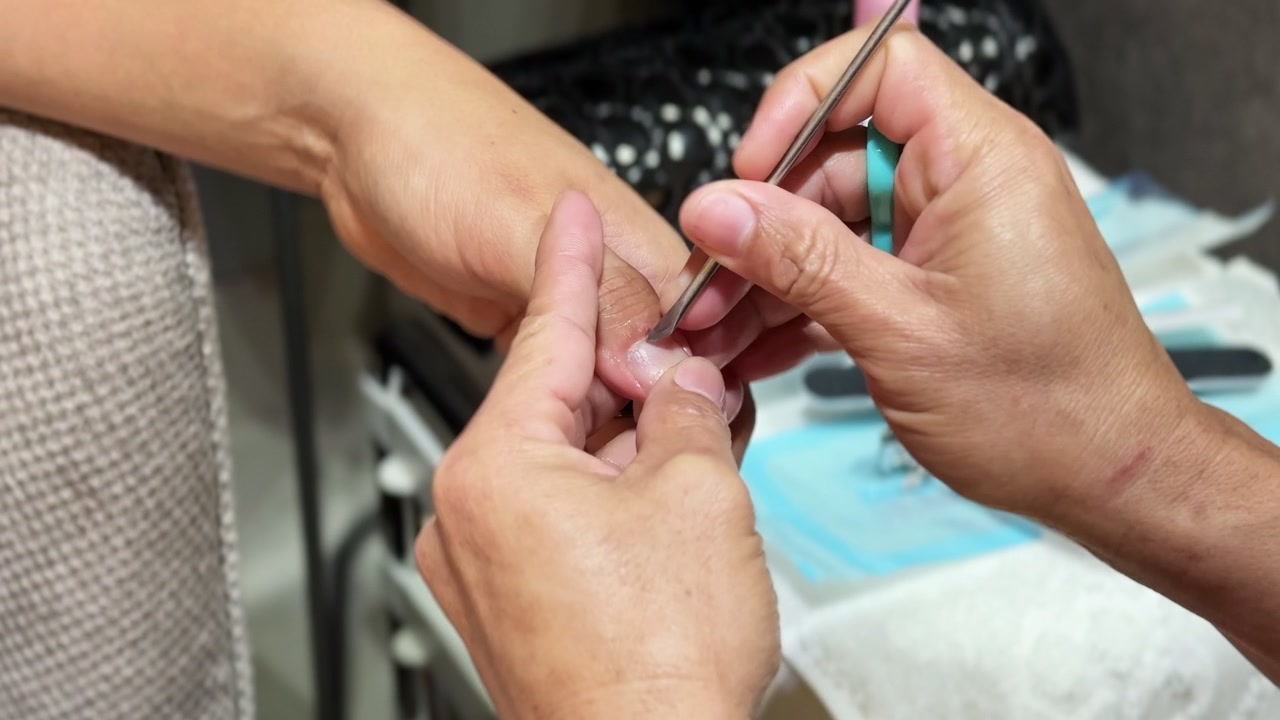 Kapalı bir güzellik salonunda, bir manikürcünün metal bir itme aletiyle müşterinin tırnak etlerini temizlemesinin yakın çekimi. Parlak yapay aydınlatma, bulanık bir ortamda profesyonel bakım uygulamasını vurguluyor.