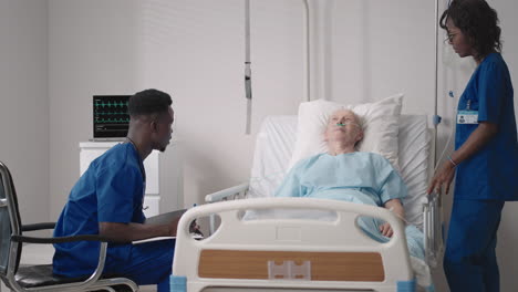 a team of african ethnic group doctors a man and a woman talk to an elderly male patient. a doctor and a nurse talk to a bedridden patient in a hospital ward